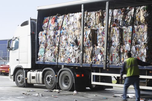 A large articulated lorry with a white cab and black trailer is parked on a paved area, fully loaded with compressed bales of mixed waste materials, including various types of paper and plastic. The bales are tightly compacted, exhibiting a rough, uneven surface with visible layers and a range of colours such as white, blue, yellow, and brown due to different printed and plain materials. The trailer’s side panels are open, revealing the dense arrangement of the waste bales stacked several layers high. In the foreground, a person wearing a reflective vest and dark trousers is walking past the vehicle, slightly blurred, indicating movement. Scattered debris is visible on the ground near the base of the trailer, and the background shows a clear sky, emphasizing the outdoor environment typical of rubbish removal or waste collection services, such as those provided by Garden Clearance Croydon in the Croydon area.