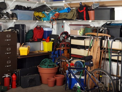 The image depicts a cluttered garage or storage room filled with various household and outdoor items. In the foreground, there is a blue bicycle leaning against a wooden shelving unit that holds plastic storage boxes, tools, and other miscellaneous objects. To the left, a large terracotta plant pot and several smaller pots are stacked on the floor, partially obscured by a coiled garden hose. Behind the pots, a black plastic storage bin, a yellow plastic container, and a red toolbox are visible on a surface, with more containers and items stacked above on a wire shelving unit. The shelving extends to the ceiling, which is partially visible, and holds additional boxes, bags, and miscellaneous objects, including some outdoor equipment and shoes. The overall scene appears disorganized, with items haphazardly placed, reflecting typical clutter in a home garage. The environment is well-lit, likely by indoor lighting, and the background features accessories and storage solutions suitable for gardening or household maintenance, consistent with typical storage areas associated with waste management and rubbish removal services in Croydon or nearby areas.
