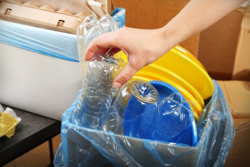 A close-up image shows a person's hand placing a clear plastic water bottle into a large transparent rubbish bag, which is filled with various colourful plastic items including yellow, blue, and possibly grey or black objects, likely lids or containers. The bag is situated on a black surface, possibly a table or worktop in an indoor or semi-indoor environment. In the background, there is a white box or bin with a partially visible blue lining or cover, and some scattered paper or packaging materials. The scene conveys a typical waste sorting or rubbish collection process, relevant to garden clearance or waste disposal services provided by Garden Clearance Croydon in the Croydon area. The focus is on the handling of recyclable plastics, with attention to detail in the textures and colours of the waste materials being loaded for removal or recycling, set against a neutral light background.