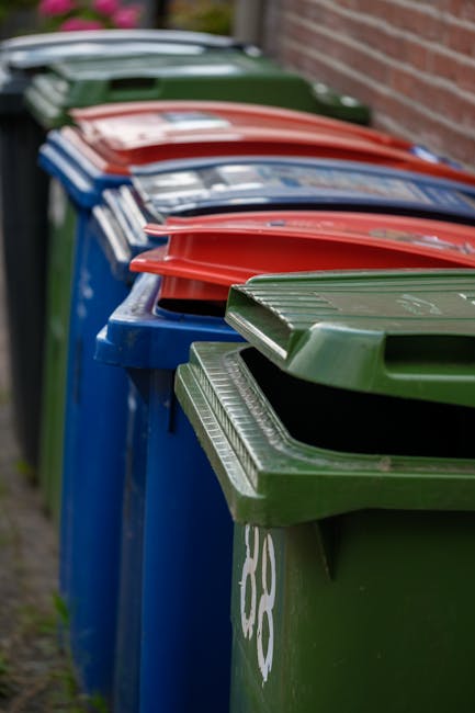 A waste collection worker wearing a high-visibility yellow and orange vest operates a red rubbish collection vehicle parked on the side of a street. The rear of the truck is open, revealing the compac