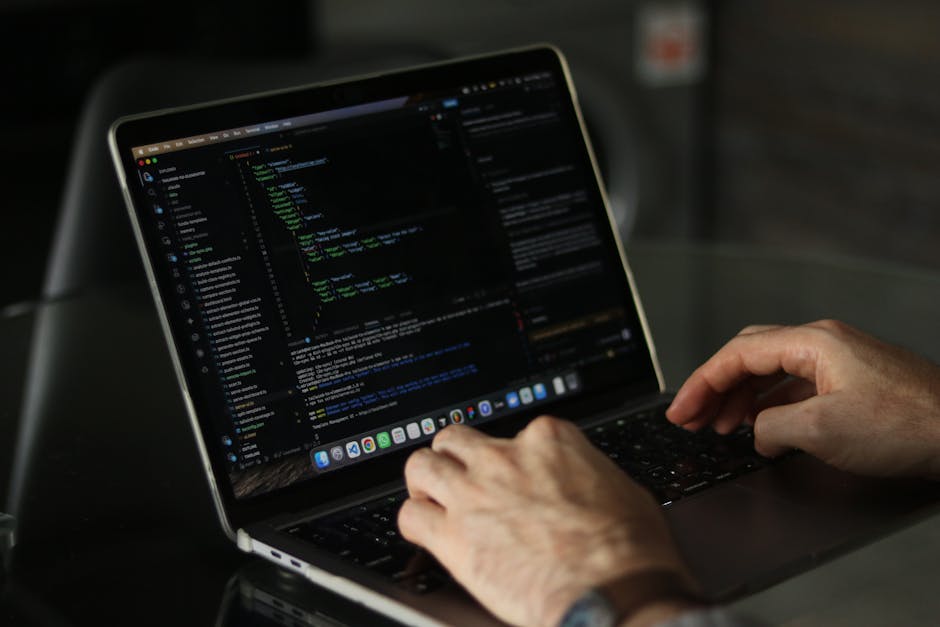 A close-up of a laptop with a black keyboard illuminated by red backlighting, displaying lines of blurred code or text on its screen, positioned on a dark surface. To the right of the laptop, there is