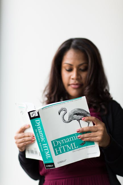 A woman with dark, wavy hair and light to medium skin tone is holding open a book titled 'Dynamic HTML' with a prominent flamingo illustration on the cover. She is standing indoors against a plain whi