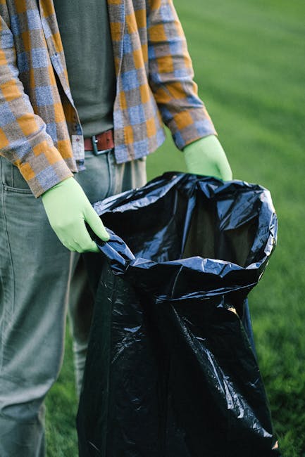 A person dressed in casual outdoor clothing, including a green long-sleeve shirt, a plaid jacket with yellow, blue, and gray tones, and a brown belt, is holding open a large black rubbish bag lined wi