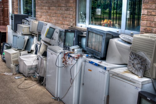 A collection of old, discarded computer monitors and CRT screens stacked outdoors on a paved area beside a brick building with a large window. The monitors vary in size, shape, and condition, with some showing visible wires and internal components, while others are mostly intact but dusty and worn. The monitors are placed directly on the ground, with some overlapping and leaning against each other. To the right, there is a metal ventilation fan and a few miscellaneous electronic parts. The scene appears to depict waste awaiting disposal or collection, with natural daylight illuminating the area, hinting at a professional rubbish removal service near Croydon. Garden Clearance Croydon manages such electronic waste and general rubbish clearance, maintaining clearances like this on driveways or storage areas.